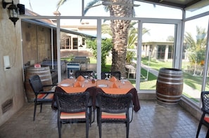 Un espacio exterior para comer con una mesa lista para cuatro personas, rodeado de sillas de mimbre con cojines, una palmera y vistas a una casa y jardín.