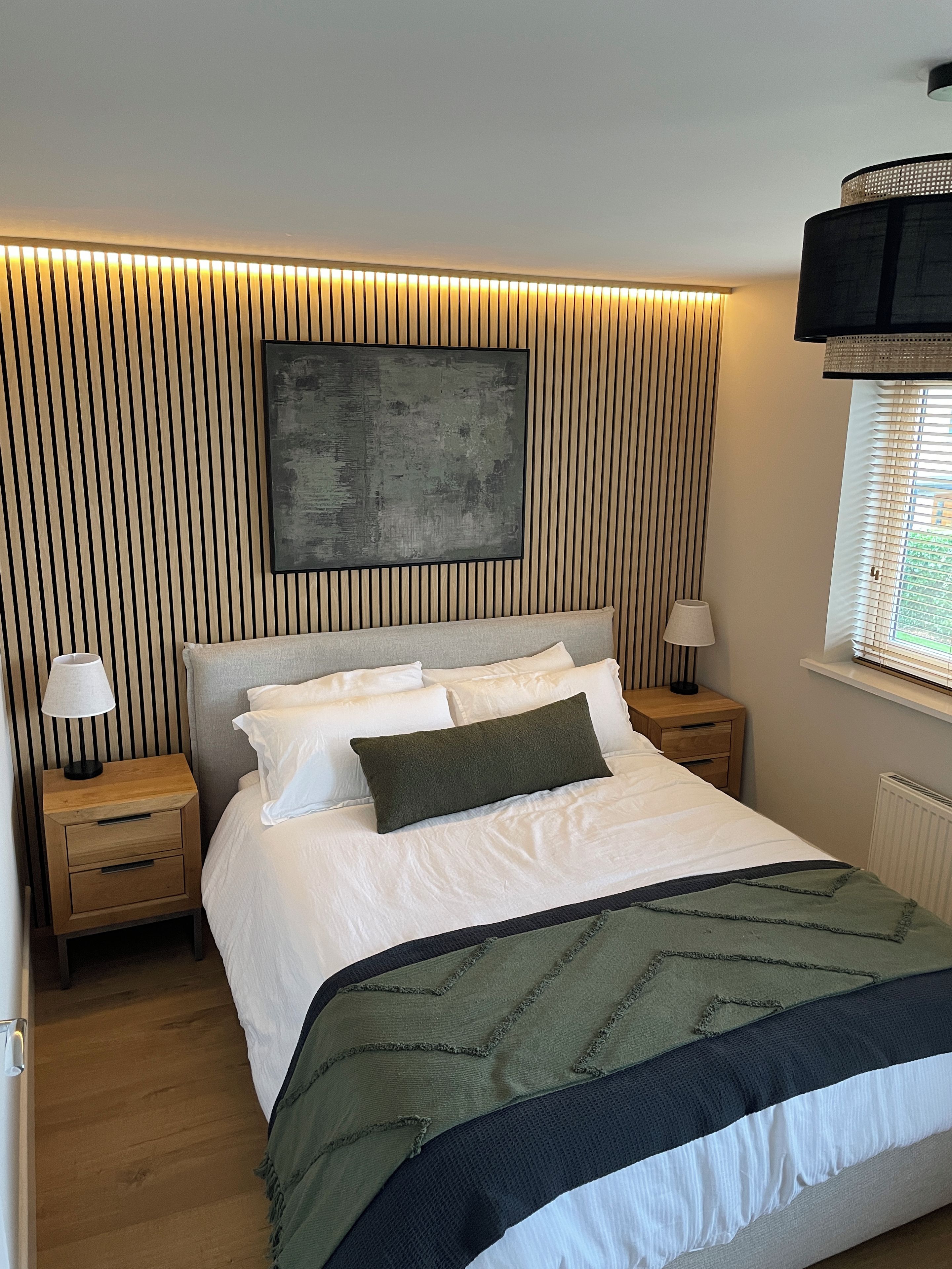 A bedroom with a bed, two bedside tables, a wall-mounted artwork, and a window with blinds.