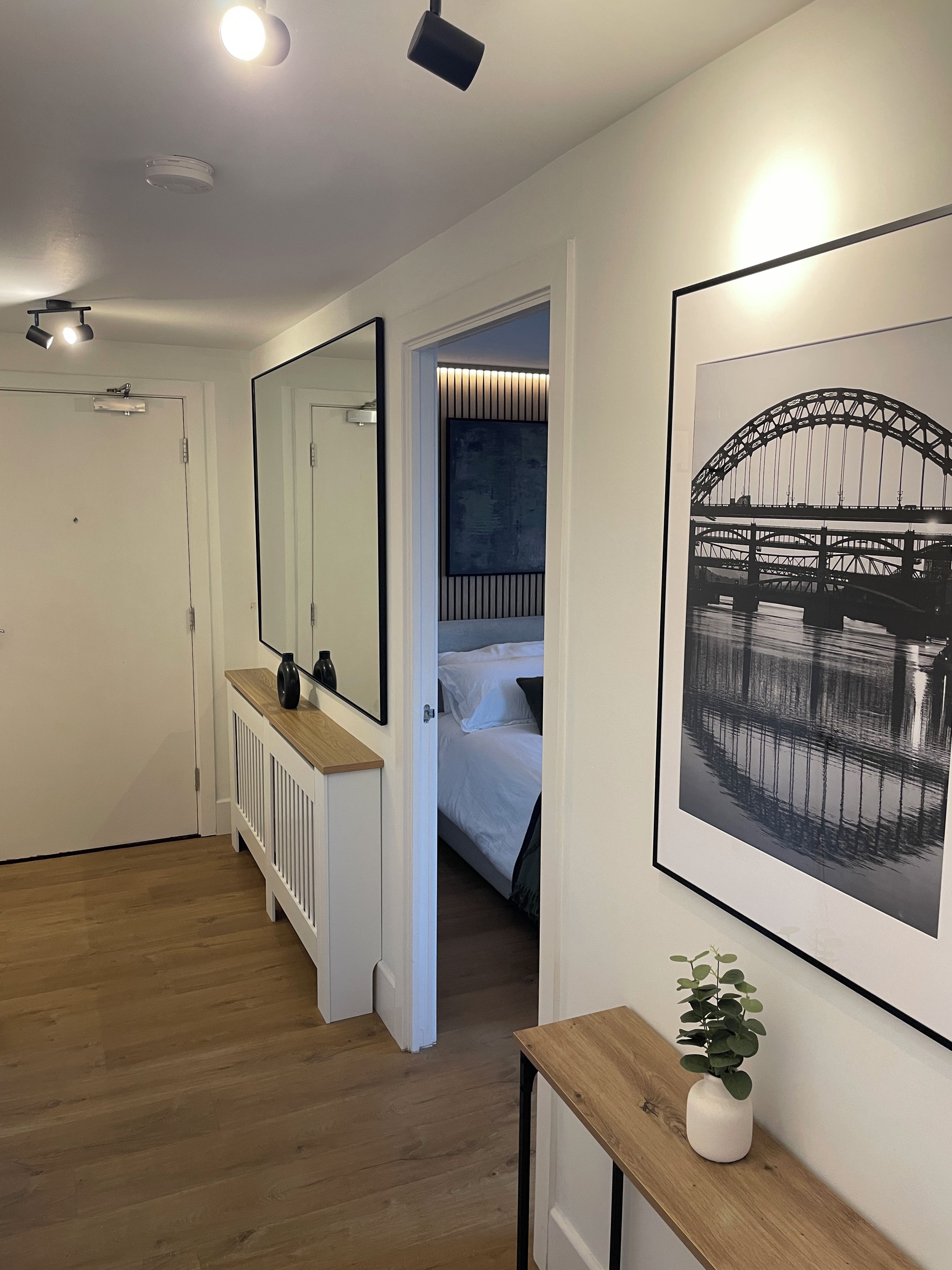 A hotel room with a wooden floor, a bedside table with a plant, a large framed picture of a bridge, and a mirror on the wall.