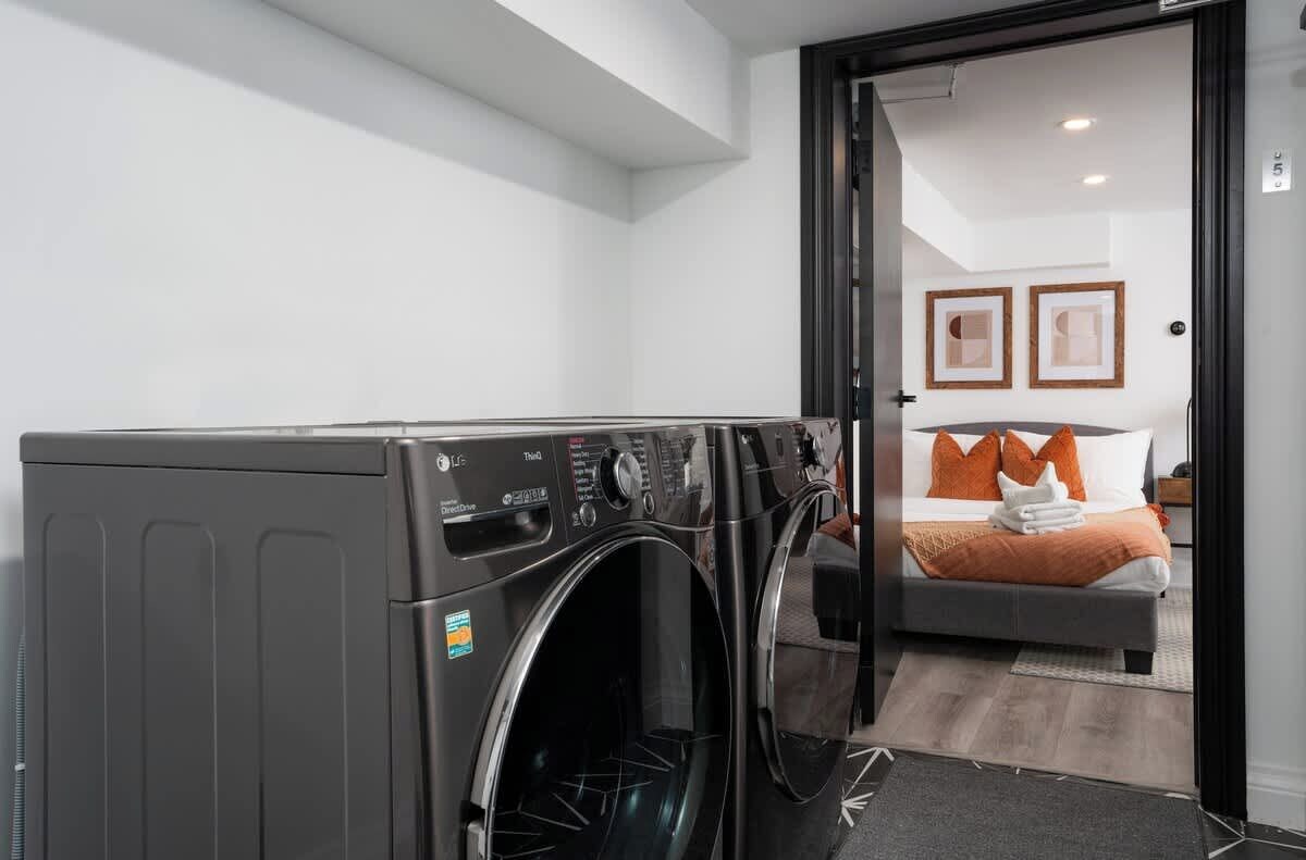 A laundry area with two front-loading washing machines, a bed with orange pillows, and a door leading to a bedroom with framed pictures on the wall.