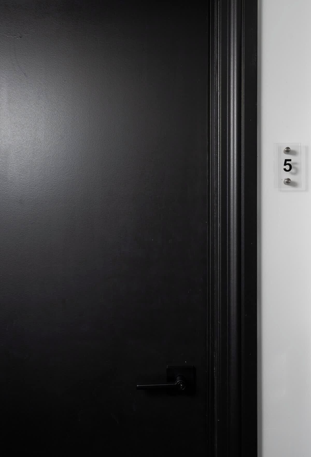 Una puerta negra con manija, una pared blanca con una placa que indica el número 5.