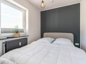 A bedroom with a bed, a window, a radiator, and a wall-mounted light fixture. - Utholm 1 Dike hut (St. Peter-Ording)
