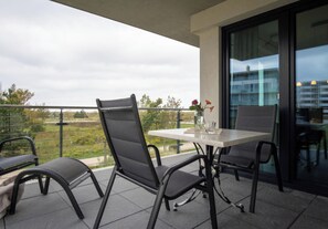 Ein Balkon mit zwei Stühlen und einem Tisch, einer Blumenvase und Blick auf ein Gebäude und Grünflächen.