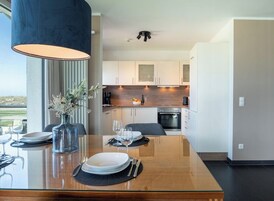 A modern dining area with a glass table, a vase with flowers, and a view of the kitchen in the background.