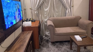 Ein Wohnzimmer mit einem Flachbildfernseher, einem beigefarbenen Sofa, einem kleinen Holztisch und einem Vorhang.