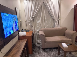 A living room with a flat-screen TV, a beige sofa, a small wooden table, and a curtain. - High Selection Serviced Apartments (Madinah)