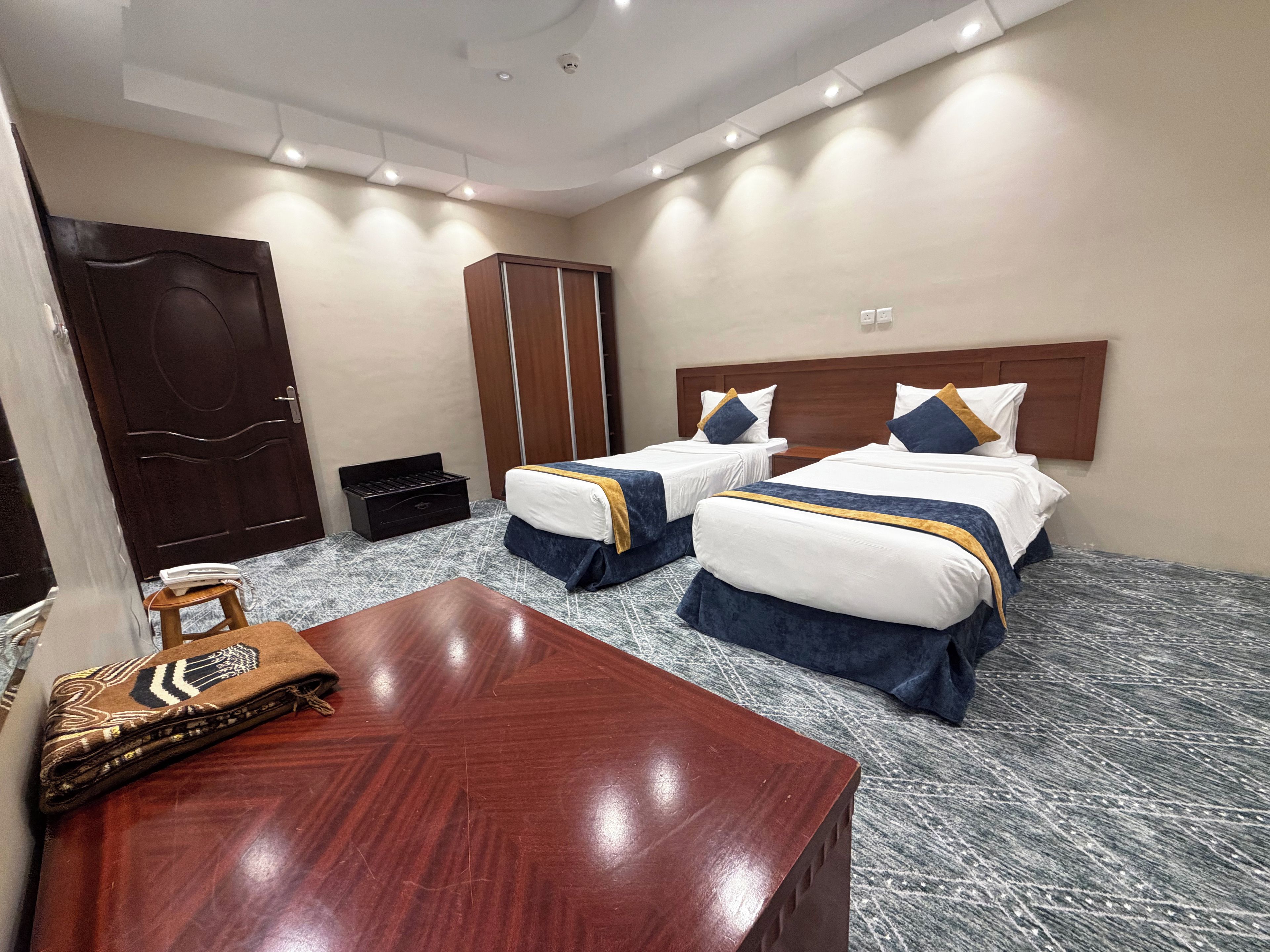 Une chambre d’hôtel avec deux lits, un bureau en bois, une armoire et une petite table recouverte d’une nappe à motifs.