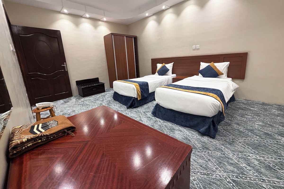 A hotel room with two beds, a wooden desk, a wardrobe, and a small table with a patterned cloth.