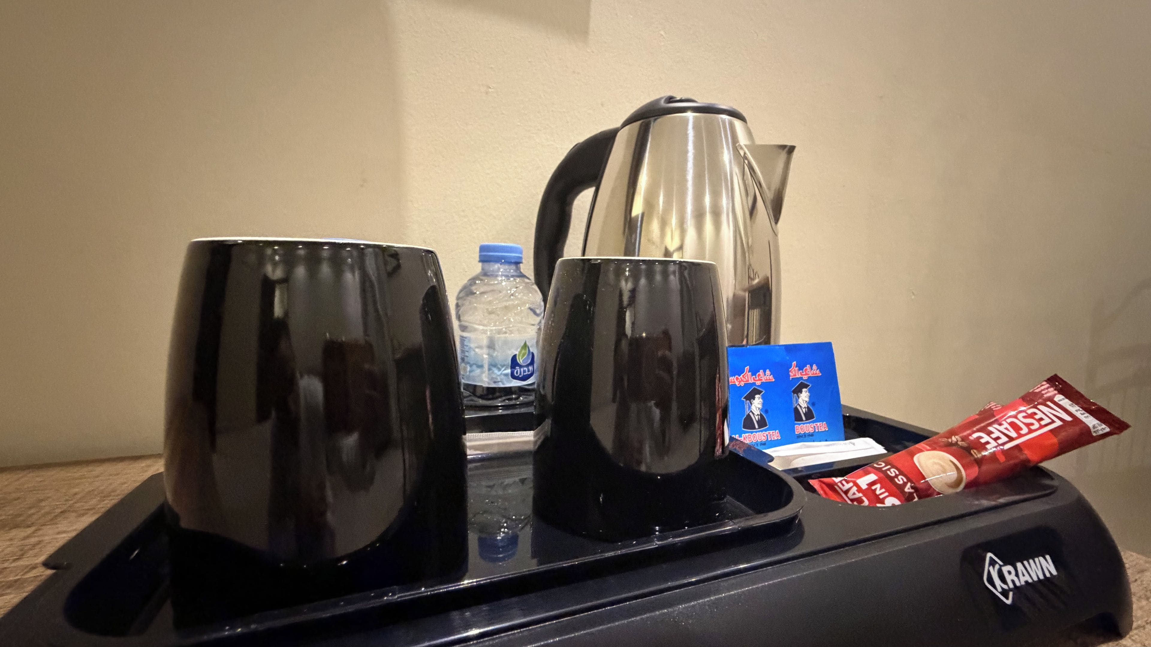A coffee station with two black mugs, a water bottle, a coffee pot, a packet of instant coffee, and a branded coffee mug.
