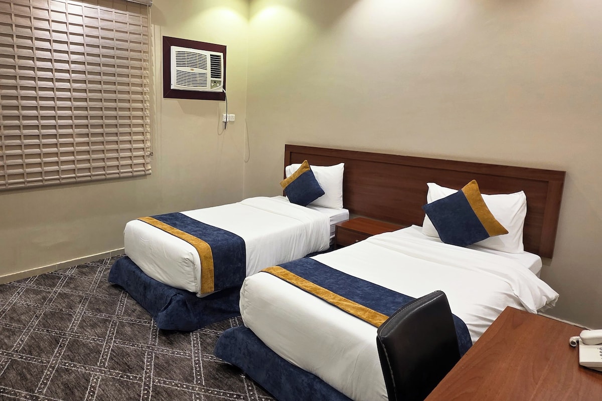 A hotel room with two beds, a wooden headboard, a bedside table, a telephone, and a window with blinds.