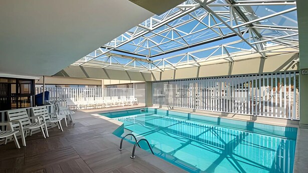 Una piscina interior con techo de cristal, rodeada de sillas blancas y suelo de madera.