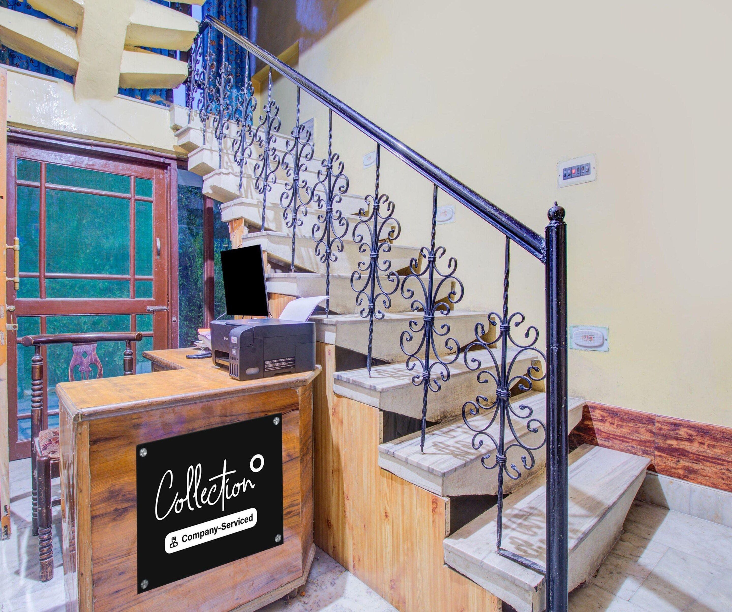 A staircase with ornate metal railings, a wooden desk with a sign reading 'Collection Company-Serviced', and a red door with a window.