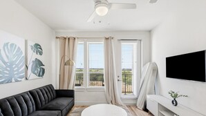 Een moderne woonkamer met een zwarte bank, een witte salontafel, een flatscreen televisie aan de muur en een groot raam met gordijnen.