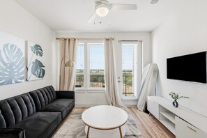 Una sala de estar moderna con un sofá negro, una mesa de centro blanca, un televisor de pantalla plana montado en la pared y un ventanal con cortinas.