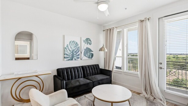 Een moderne woonkamer met een zwarte bank, een witte fauteuil en een ronde salontafel. Er is een groot raam met gordijnen, een plafondventilator en kunst aan de muur.