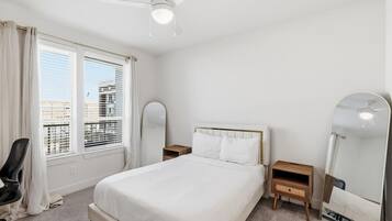 Une chambre d’hôtel équipée d’un lit, d’un bureau, d’une chaise, d’un ventilateur de plafond et d’une grande fenêtre avec des stores.