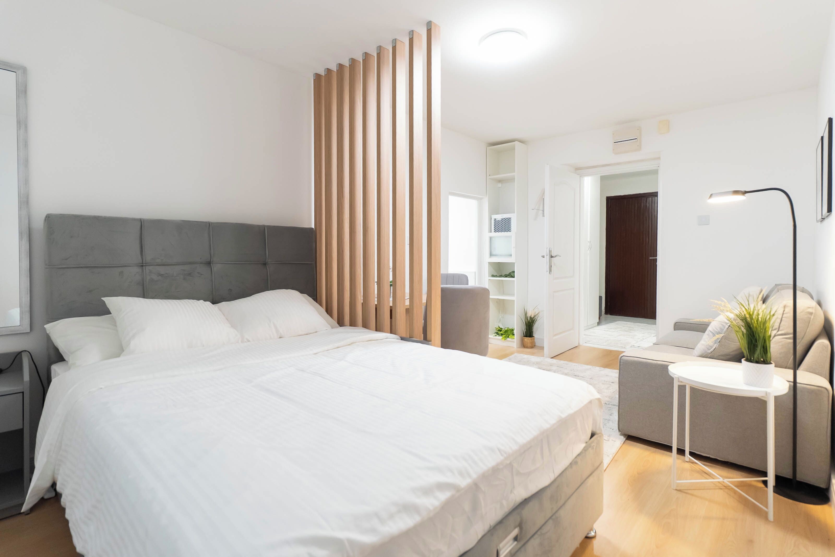 A modern bedroom with a large bed, a grey headboard, a wooden partition, a bedside table, a lamp, and a small seating area with a plant.