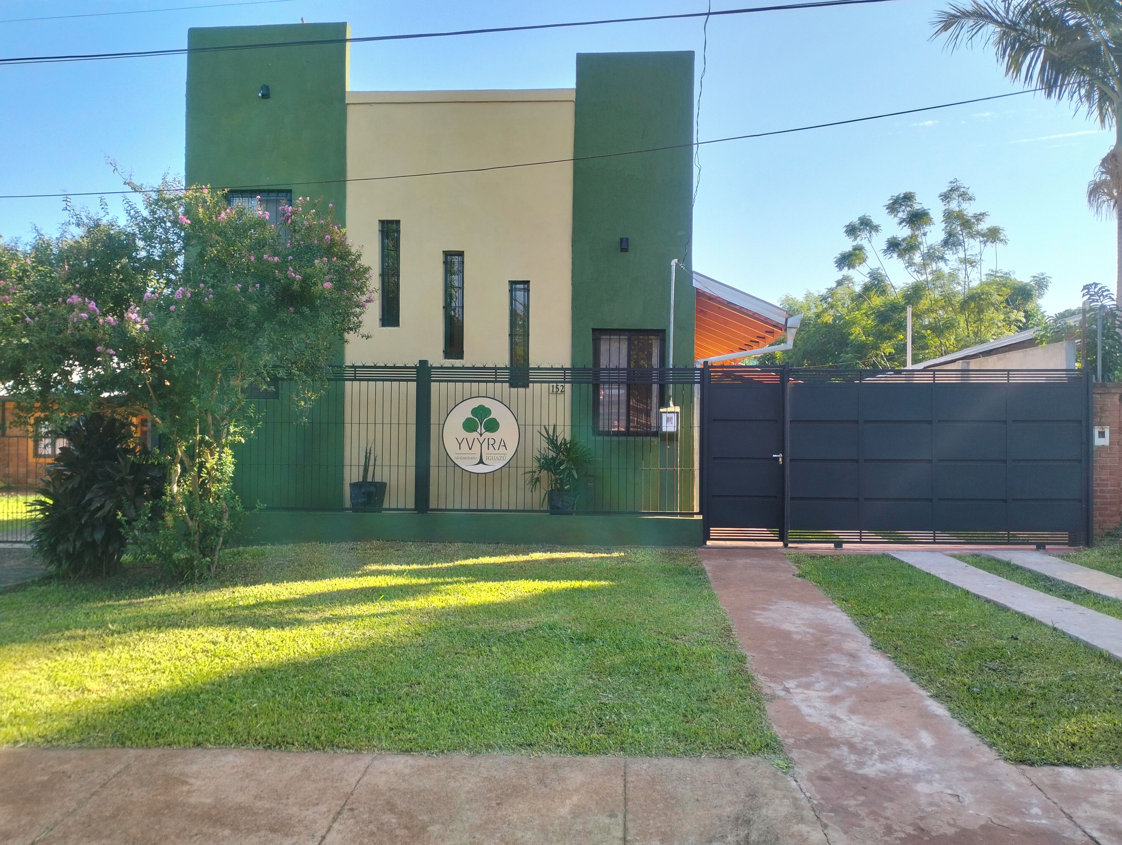 Una propiedad con acceso restringido, fachada verde y beige, un logotipo con un árbol y un camino de entrada pavimentado.