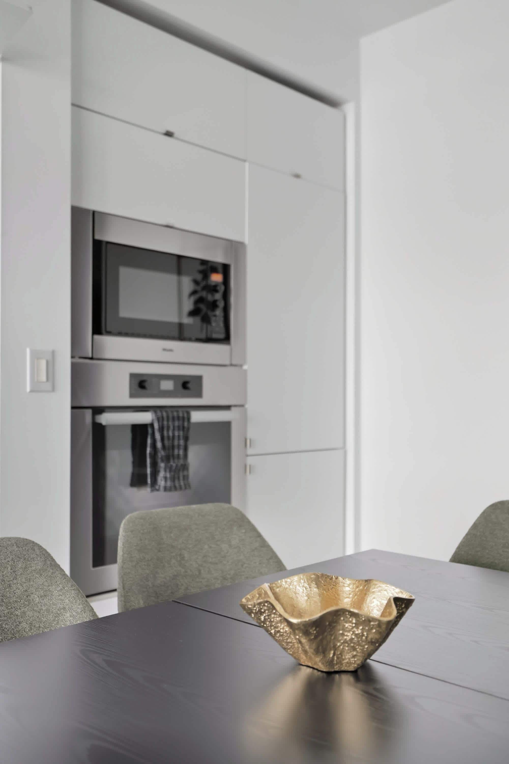 A modern kitchen with stainless steel appliances, a dining table with a unique gold bowl centerpiece, and grey upholstered chairs.