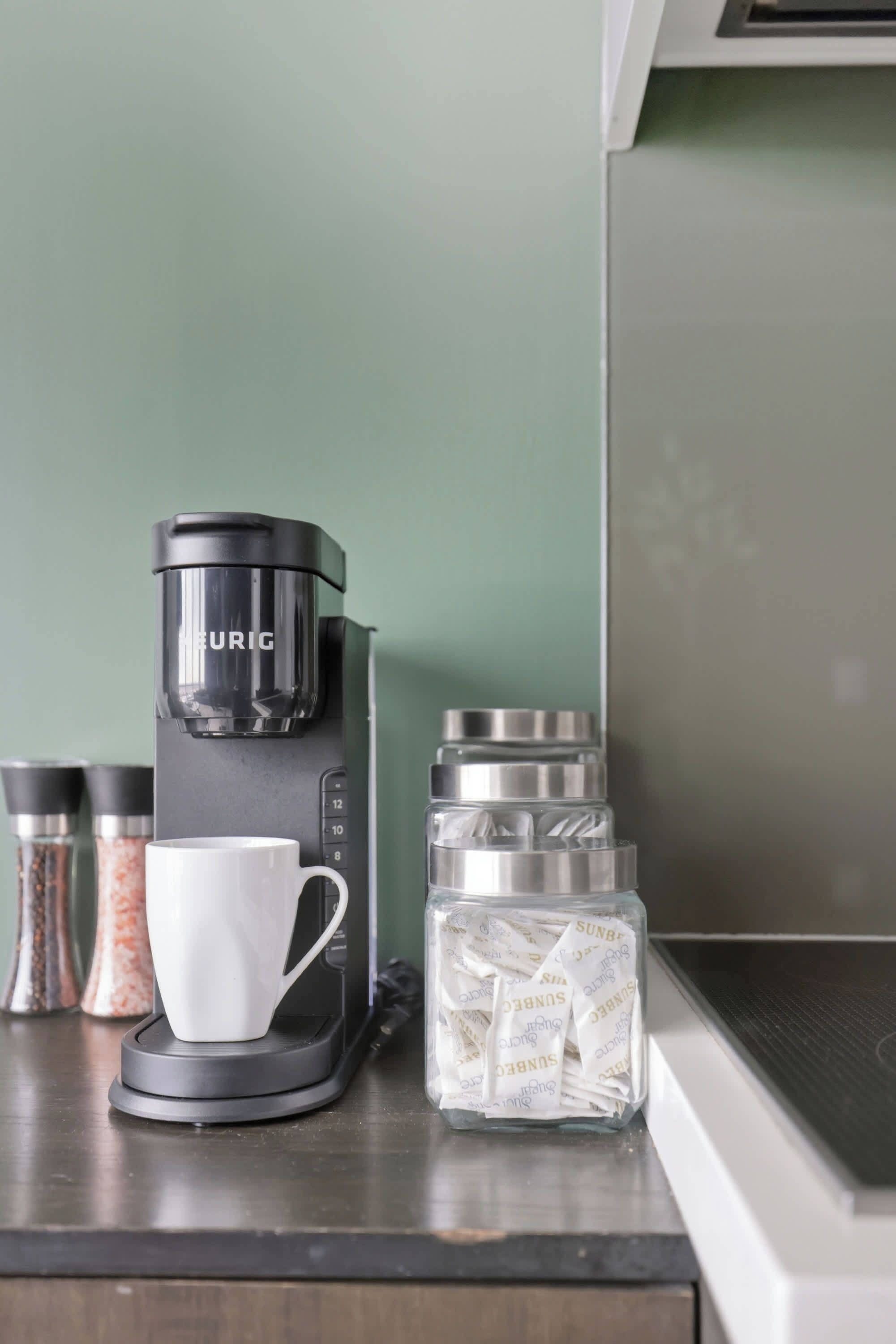 Una encimera de cocina con una cafetera negra, una taza blanca y dos frascos de especias.