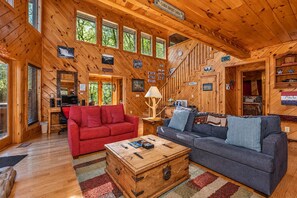 Interior de una cabaña con un sofá rojo, un sofá azul, un cofre de madera y una escalera.