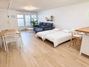 Et moderne, godt oplyst rum med trægulv, en grå sofa, en seng med hvide sengetæpper, et lille rundt bord og en hvid stol.