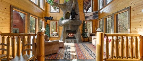 A log cabin interior with a fireplace, a large tree in the center, and a patterned rug.