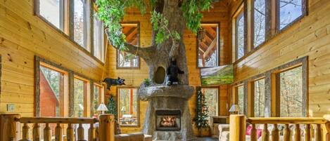 Interior de una cabaña de troncos con un gran árbol como centro, una chimenea y un rincón de estar acogedor.