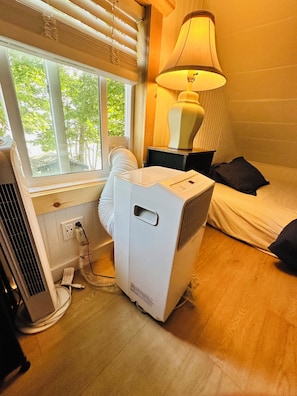 A room with a bed, a standing heater, a window with blinds, and a lamp.