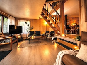 A cozy living room with a wooden staircase, a flat-screen TV, and comfortable seating.