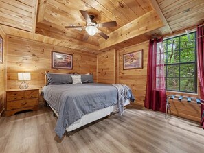 A wooden cabin bedroom with a bed, a nightstand, a ceiling fan, and a window with curtains. - Peaceful 3BR w/Hot Tub*Theater*Game Room*Privacy! (Sevierville)