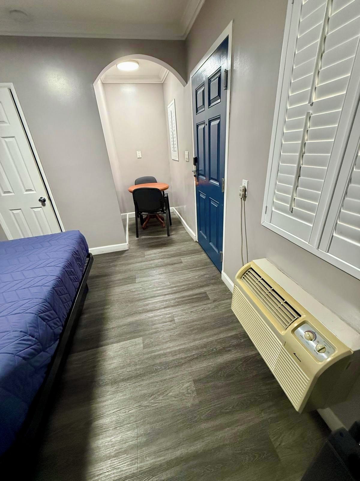 A narrow hallway with a bed on the left, a chair in the center, and a door at the end.