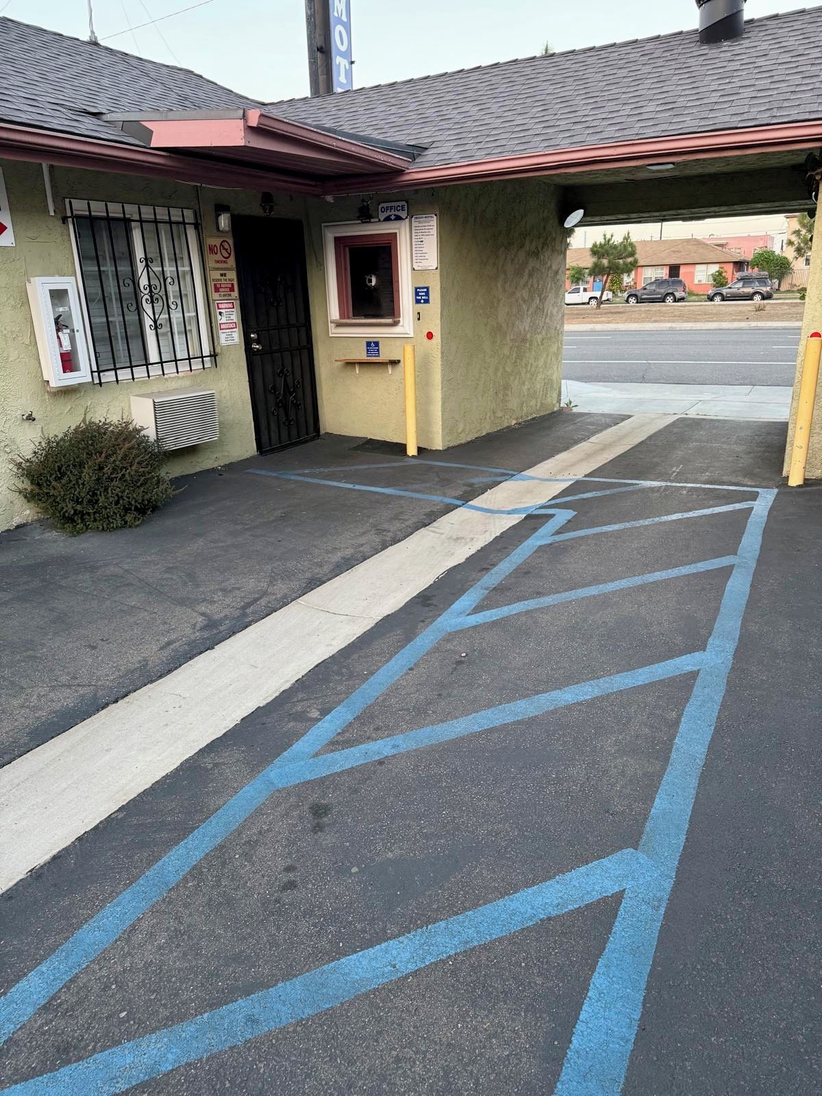 Un edificio con toldo rojo, puerta negra y ventanal con reja de metal. Se observa un espacio de estacionamiento azul con líneas diagonales.