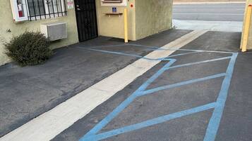Un edificio con toldo rojo, puerta negra y ventanal con reja de metal. Se observa un espacio de estacionamiento azul con líneas diagonales.