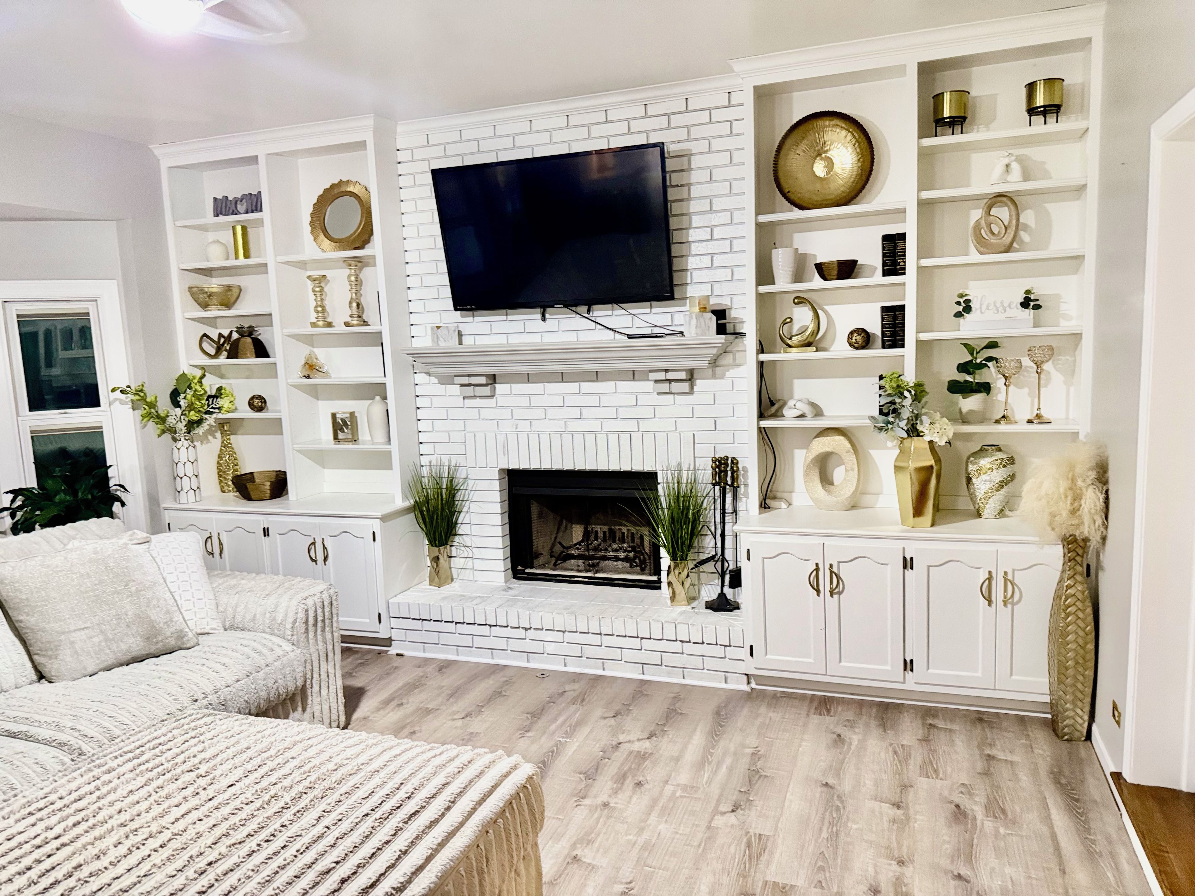 Un salon doté d’une cheminée en briques blanches, d’un téléviseur à écran plat fixé au-dessus, et d’une étagère blanche agrémentée d’objets décoratifs.