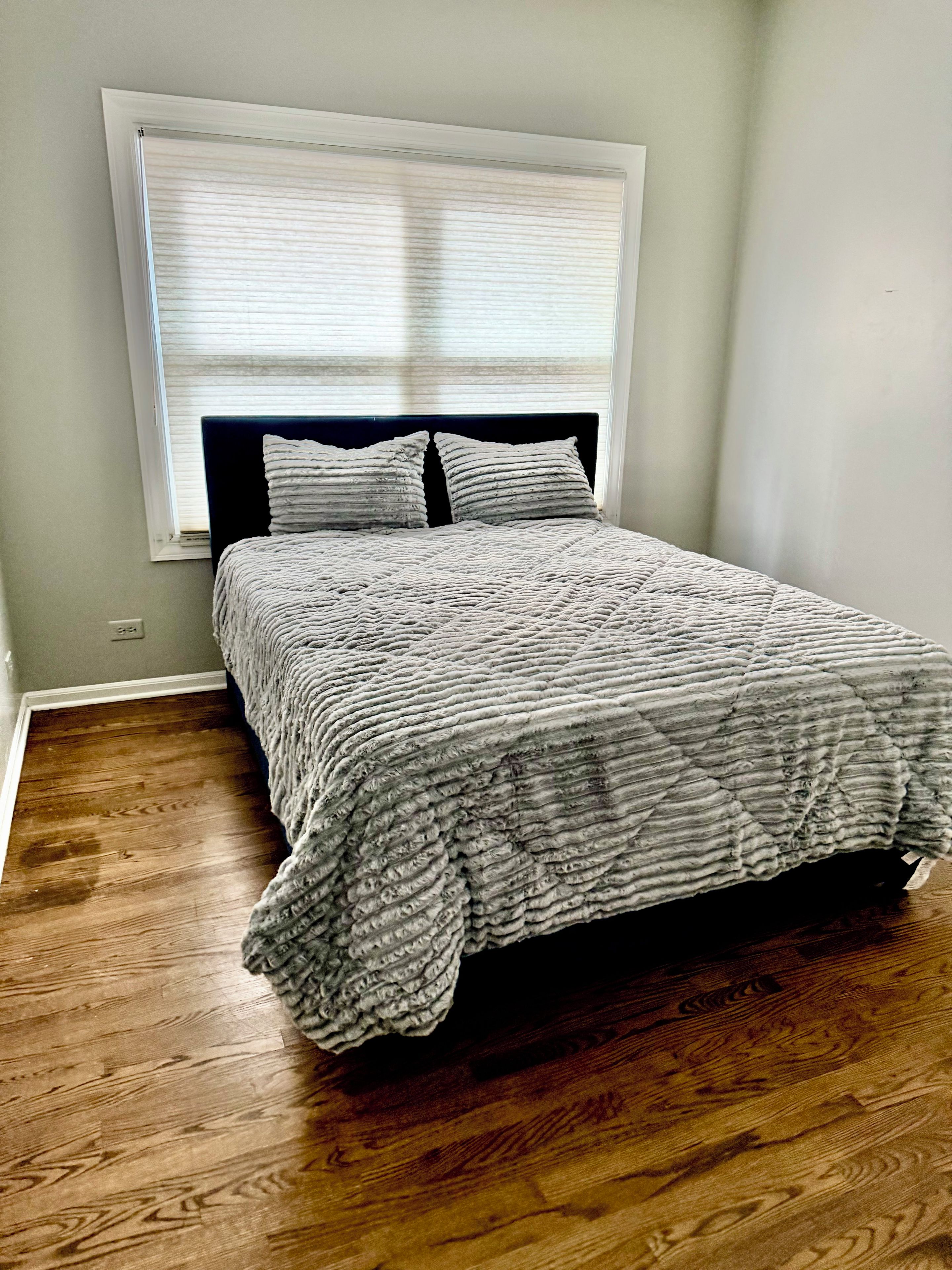 A bedroom with a bed, a window with blinds, and a wooden floor.