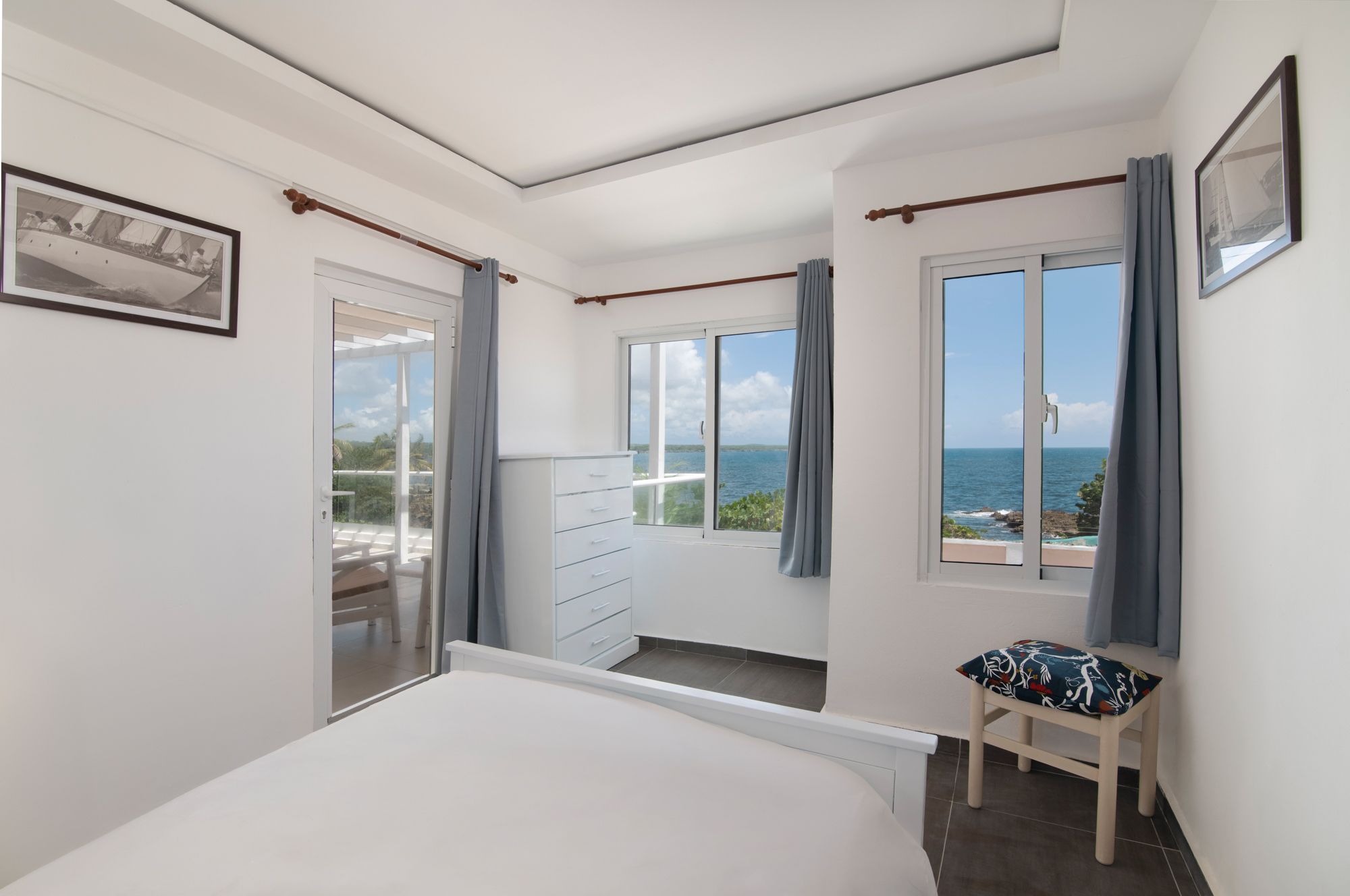 A bedroom with a bed, a chair, a dresser, and a window with a view of the sea.