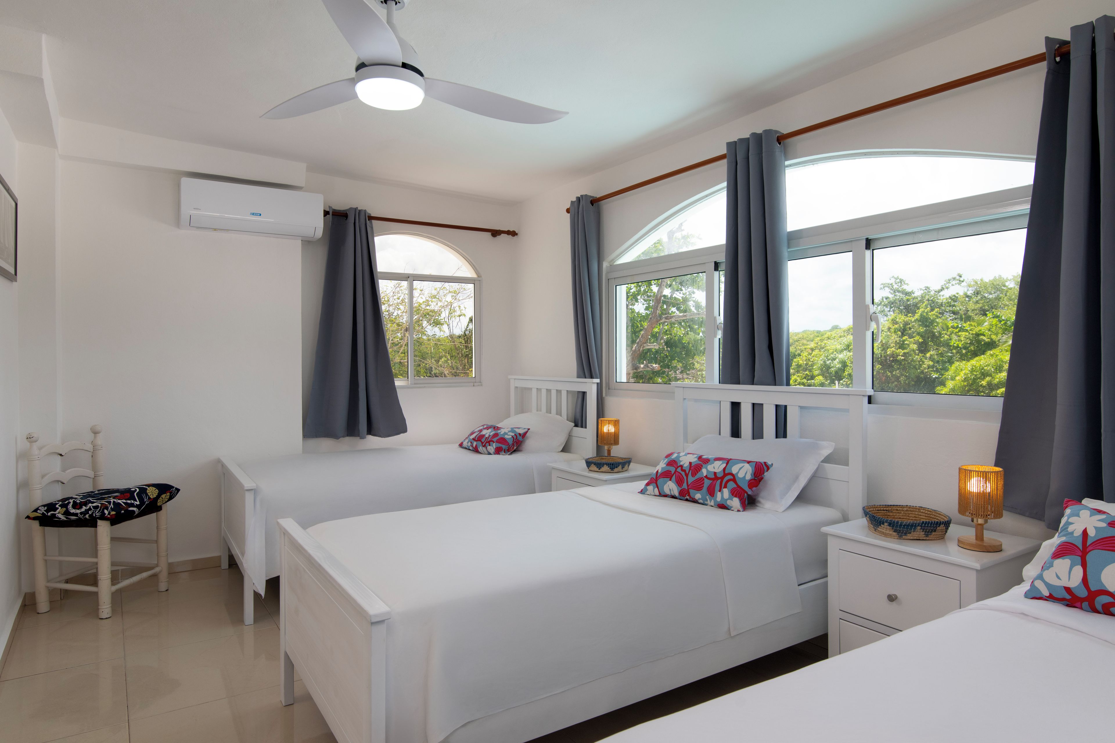 Une chambre d’hôtel avec deux lits, un ventilateur de plafond, un appareil de climatisation, une fenêtre avec des rideaux, une table de chevet avec une lampe et une chaise.