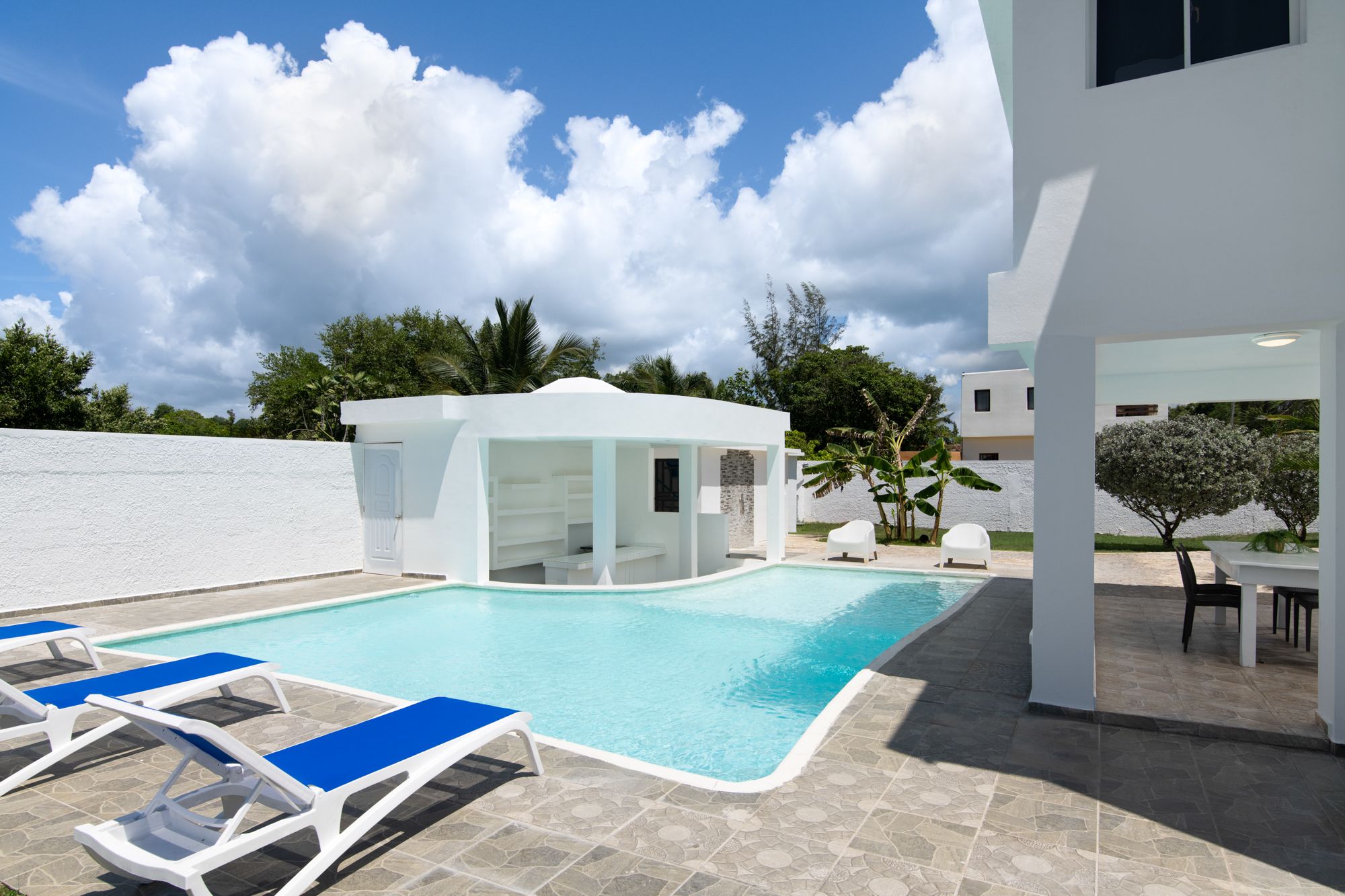 A modern outdoor pool area with lounge chairs, a covered seating area, and a white building with a door.