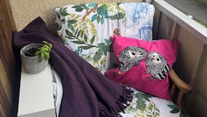 Parvekkeella on pieni pöytä, ruukkuistutettu kasvi sekä tuoli, jossa on kukalliset koristetyynyt ja vaaleanpunainen koristetyyny.