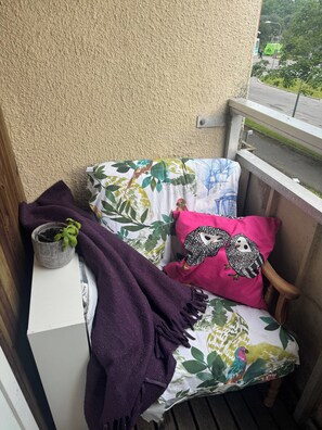 Un balcón con una pequeña mesa, una planta en maceta y una silla con cojines estampados y un cojín rosa.