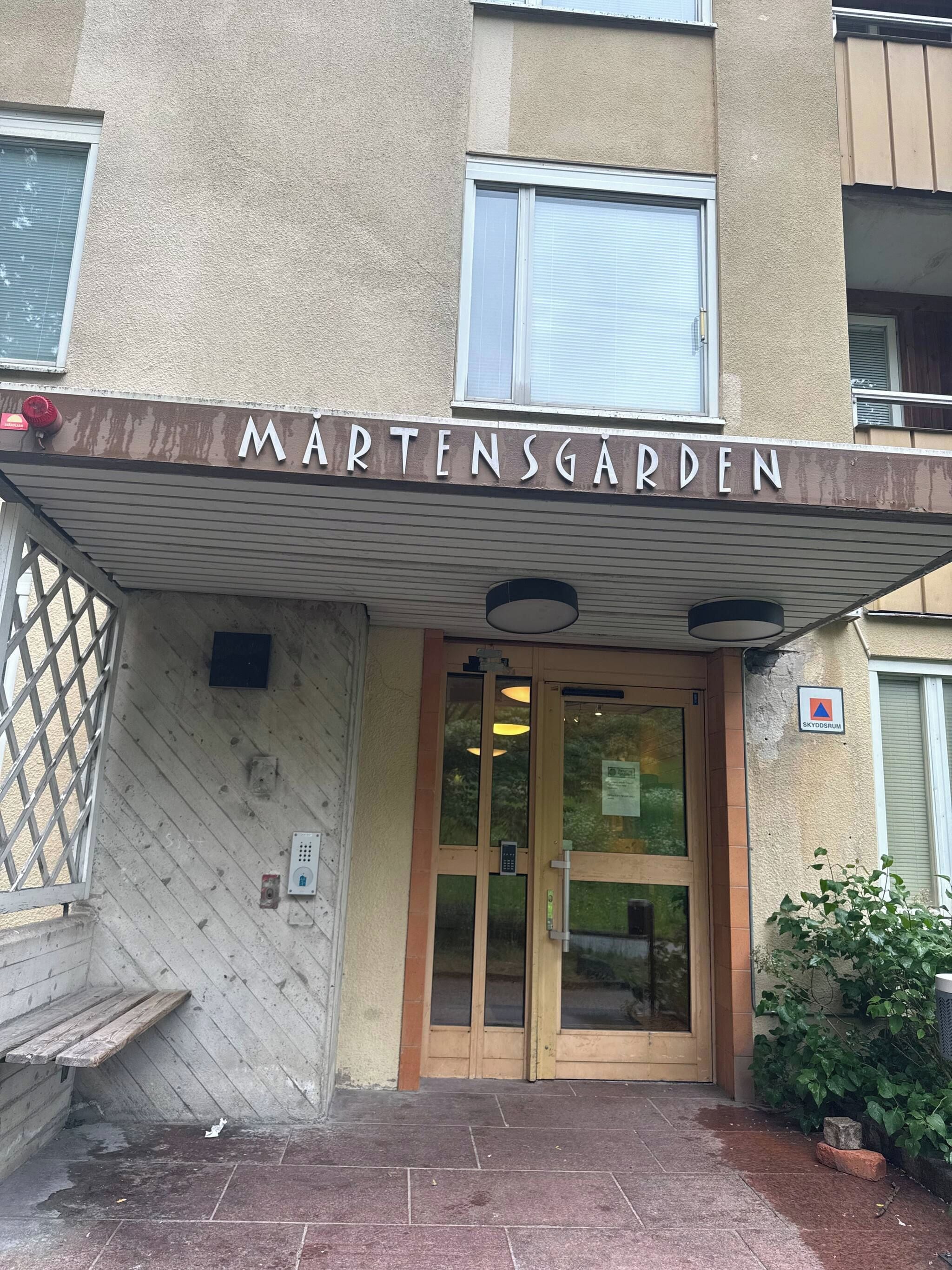 The entrance to a building named MARTENSGARDEN with a covered entrance, a glass door, and a small outdoor area with a bench.