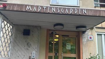 The entrance to a building named MARTENSGARDEN with a covered entrance, a glass door, and a small outdoor area with a bench.