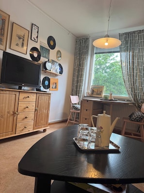 Habitación con televisor, mueble de madera, mesa con tetera y vasos, y ventana con cortinas.