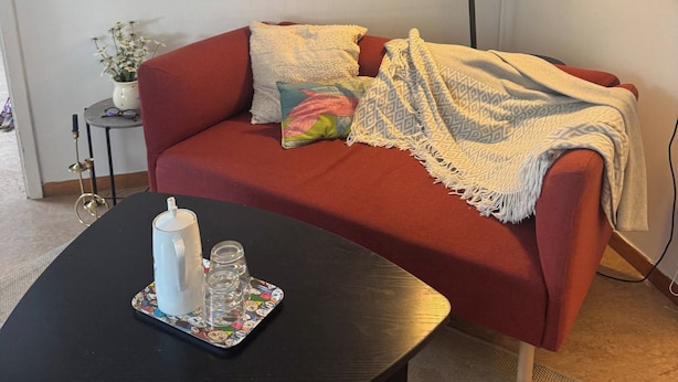 Huoneessa on punainen sohva, musta sohvapöytä, jonka päällä on muki ja laseja, lattiavalaisin sekä pieni sivupöytä, jonka päällä on kasvi.