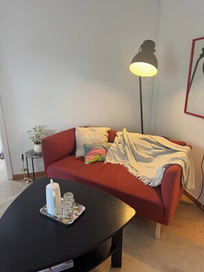 Una habitación con un sofá rojo, una mesa de centro negra con una taza y vasos, una lámpara de pie y una mesita auxiliar con una planta.