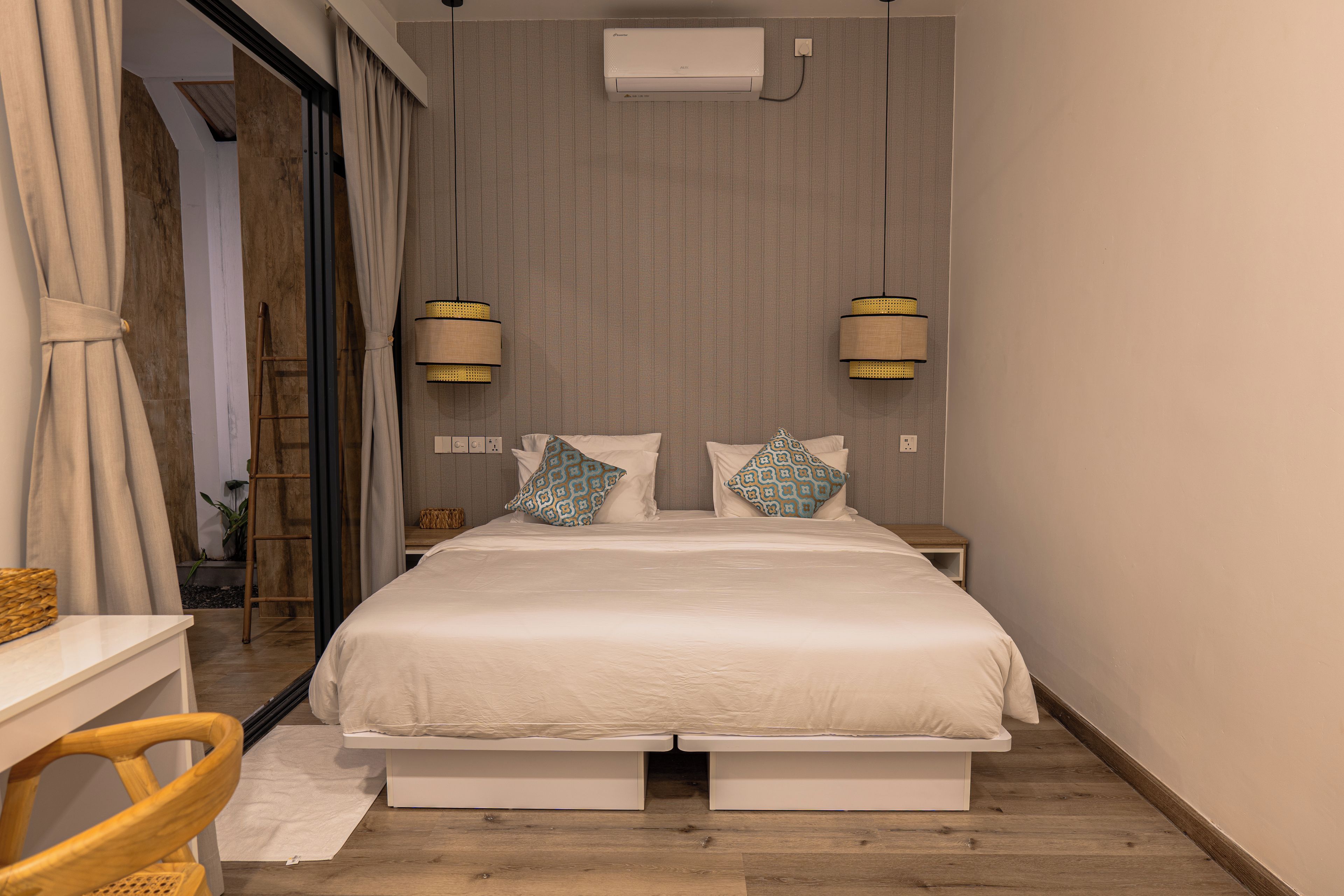 A bedroom with a bed, two bedside lamps, a wall-mounted air conditioner, and a wooden floor.