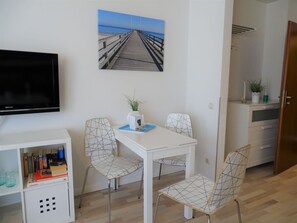 A room with a white table, two chairs, a flat-screen TV, a bookshelf, and a painting of a pier. - Beach House Villa 9 (Dahme)
