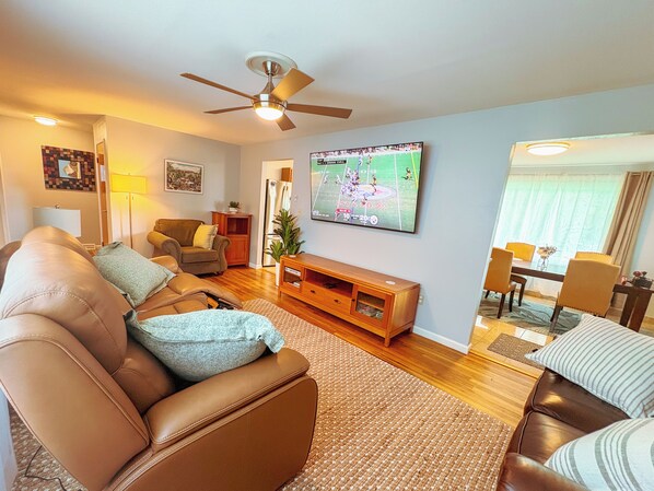 Un salon avec un canapé en cuir marron, un meuble de rangement en bois, un ventilateur de plafond et un téléviseur à écran plat diffusant un match de sport.