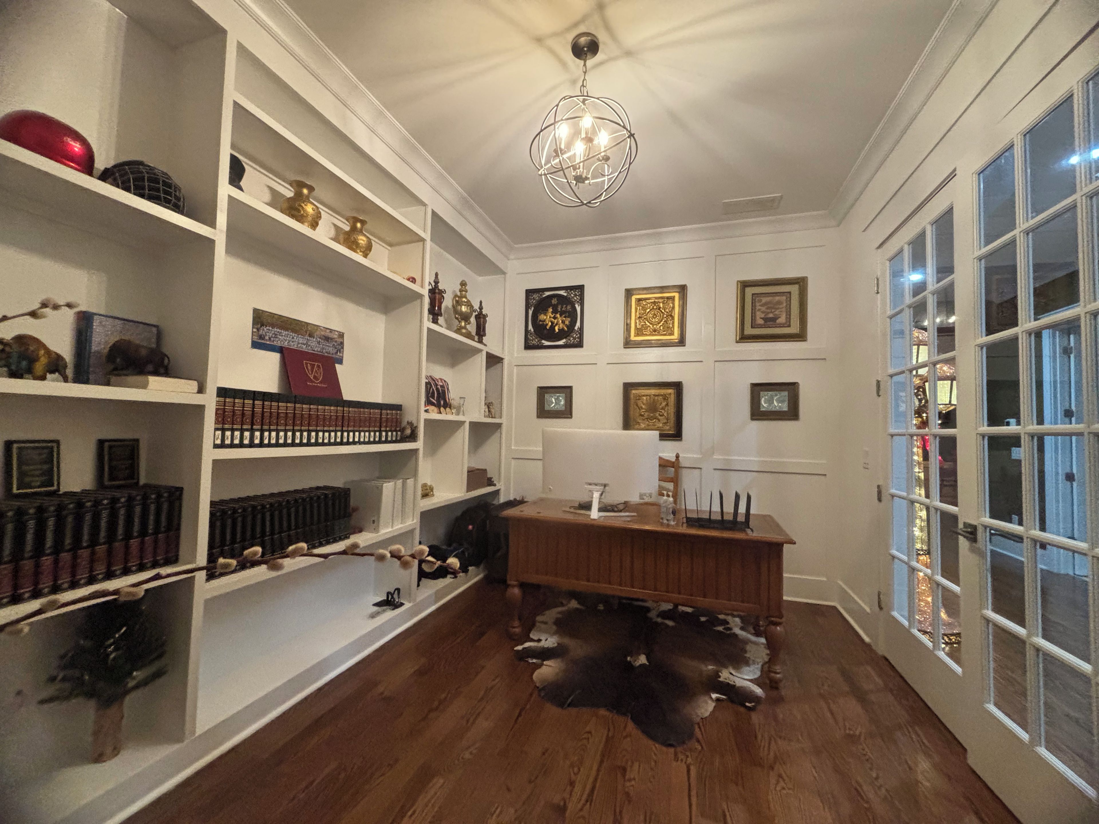 A room with white walls, wooden flooring, a desk with a leather rug, shelves with books and decorative items, and a chandelier.